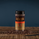 A jar of Bourbon Barrel Foods Bourbon Smoked Cacao Nibs sits on a rustic wooden surface, some nibs scattered around. The brown and gold label highlights Louisville, Kentucky—ideal for baking or snacking.