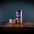 Two containers of Bourbon Barrel Foods Bourbon Smoked Chef's Blend seasoning—one small metal tin and one tall plastic jar—sit on a wooden surface with some spice blend scattered nearby, set against a dark blue background.