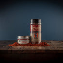 Two containers of Bourbon Barrel Foods Bourbon Smoked Chili Powder—one tin and one plastic shaker—sit on a wooden surface with red seasoning scattered in front, against a dark blue background.