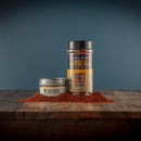 A jar and a tin of Bourbon Barrel Foods Bourbon Smoked Chili & Coffee Rub rest on a wooden surface, smoky seasoning scattered in front, showcasing this bold BBQ rub by Bourbon Barrel Foods against a plain blue background.