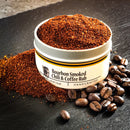 A tin of Bourbon Barrel Foods Bourbon Smoked Chili & Coffee Rub sits on a dark surface, filled with reddish-brown smoky seasoning. Some BBQ rub is spilled nearby, with whole coffee beans scattered around the tin.