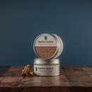 Two tins of Bourbon Barrel Foods Bourbon Smoked Cinnamon Sugar are stacked on a rustic wooden surface with two cinnamon sticks in front, set against a plain blue background.