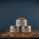 Three tins of Bourbon Barrel Foods Bourbon Smoked Cinnamon Sugar are stacked on a rustic wooden surface, with cinnamon sugar spilling from the top and scattered at the base, set against a plain blue background.