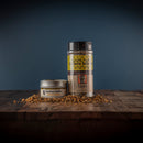 Two containers of Bourbon Barrel Foods Bourbon Smoked Citrus Pepper—one tin and one plastic shaker—sit on a wooden surface with spilled seasoning, ideal for bold grilled meats, set against a dark blue background.