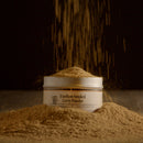A tin of Bourbon Barrel Foods Bourbon Smoked Curry Powder rests on a mound of rich, smoky curry spice, as more golden powder falls above against a dark background.