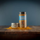 Two containers of Bourbon Barrel Foods Bourbon Smoked Curry Powder, a small tin and a tall shaker, rest on wood surrounded by golden curry powder and smoky spice blend, with powder scattered against a blue backdrop.