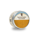 Bourbon Barrel Foods Bourbon Smoked Curry Powder comes in a round tin with a clear lid, displaying its yellow-brown curry spice blend—offering a smoky twist—on a light background.