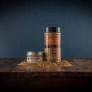 Two jars of Bourbon Barrel Foods Bourbon Smoked Garlic Salt rest on a wooden surface, with some of the savory, smoky salt sprinkled nearby. A dark gradient background highlights this flavorful Bourbon Barrel Foods seasoning.