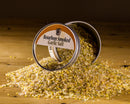 A tin of Bourbon Barrel Foods Bourbon Smoked Garlic Salt rests on its side, scattering yellow and white smoky salt granules from Bourbon Barrel Foods onto a wooden surface.