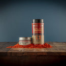 Two containers of Bourbon Barrel Foods Bourbon Smoked Paprika—a small tin and a tall jar—rest on wood with a pile of vibrant red paprika in front, set against a dark blue background.