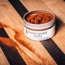 An open tin of Bourbon Barrel Foods Bourbon Smoked Paprika rests on a wooden surface, with some of the rich, bourbon barrel-smoked spice spilled out beside it.