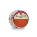 A round tin of Bourbon Barrel Foods Bourbon Smoked Paprika, featuring a maroon and tan label and partially removed metal lid, contains reddish-orange paprika smoked in bourbon barrels.