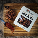 Bourbon Barrel Foods Bourbon Smoked Pecans sit on a wooden board with pecans and a metal scoop of red spice—a smoky, irresistible snack for charcuterie boards. The clear window on the box reveals the delicious pecans inside.