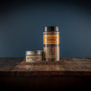 Two containers of Bourbon Barrel Foods Bourbon Smoked Pepper—one metal tin, one clear shaker with matching brown and yellow labels—rest on a rustic wooden surface, surrounded by scattered smoked Malabar pepper.