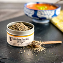 A tin of Bourbon Barrel Foods Bourbon Smoked Pepper—with some pepper spilled and a small wooden spoon—rests on a black surface; a blurred bowl of red soup and a yellow napkin appear in the background.
