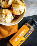 A bowl of vanilla ice cream topped with Bourbon Barrel Foods Bourbon Smoked Sea Salt Caramel Sauce sits on a dark surface beside its bottle, with an orange cloth underneath the bowl.