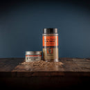Two containers of Bourbon Barrel Foods Bourbon Smoked Sea Salt, a small tin and a tall plastic jar, sit on rustic wood with scattered seasoning, set against a dark blue background.