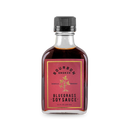 A clear glass bottle with a black cap contains dark liquid. The label reads Bourbon Barrel Foods Bourbon Smoked Soy Sauce, features gold and red accents, decorative text, a plant illustration, and notes it's bourbon barrel aged and non-GMO.