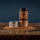 Two containers of Bourbon Barrel Foods Bourbon Smoked Sugar, a small tin and a large jar, sit on a rustic wooden surface with sugar granules scattered around them, set against a dark blue background.