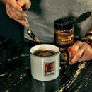 Someone adds a spoonful of Bourbon Barrel Foods Bourbon Smoked Sugar to coffee in a mug with a Bourbon logo, next to an open container of the same sugar on a dark countertop.