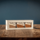 The Bourbon Barrel Foods Bourbon Smoked Sugars Trio—Orange, Vanilla & Cinnamon—comes in a white rectangular box with three jars, shown resting on wood with a blue background.