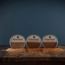 The Bourbon Barrel Foods Bourbon Smoked Sugars Trio—Orange, Vanilla & Cinnamon—sits on a rustic wood surface with gourmet sugars sprinkled in front, against a dark blue background.