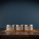 Three tins of Bourbon Barrel Foods Bourbon Smoked Sugars Trio—Orange, Vanilla & Cinnamon—rest on a wooden surface with scattered granules, set against a dark blue background.