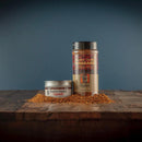 Two containers of Bourbon Barrel Foods Bourbon Smoked Togarashi sit on a wooden surface with piles of the reddish Japanese-inspired spice around them, set against a dark blue background.