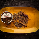 A yellow rectangular plate displays a tin of Bourbon Barrel Foods Bourbon Smoked Togarashi, a Japanese-inspired seasoning blend, next to mixed spices, a dried red chili pepper, and a cinnamon stick on a dark wooden surface.