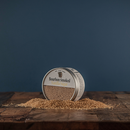 A small tin of Bourbon Barrel Foods Bourbon Smoked Vanilla Sugar is tipped over on a wooden surface, spilling light brown, Madagascar vanilla–infused sugar granules against a plain dark blue background.