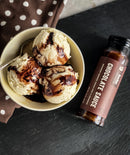 A bowl with three scoops of vanilla ice cream drizzled with Bourbon Barrel Foods Chocolate Sauce – Bourbon Smoked Cacao Nibs & Bourbon sits beside the bottle on a dark surface with a brown polka dot cloth nearby.