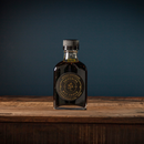 A glass bottle of Bourbon Barrel Foods Imperial Double Fermented Bluegrass Soy Sauce, topped with a black cap, rests on rustic wood before a dark blue backdrop. Its black and gold label showcases the brand’s superior quality.