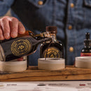 Wearing a denim shirt, someone pours Bourbon Barrel Foods' Imperial Double Fermented Bluegrass Soy Sauce from its labeled glass bottle into a small dish, with more bottles and dishes on a wooden tray in front of them.
