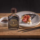 A bottle of Bourbon Barrel Foods Imperial Double Fermented Bluegrass Soy Sauce rests on a wooden board beside a soy sauce dish, sushi plate, and black chopsticks set against a dark background.