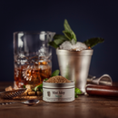 A tin of Bourbon Barrel Foods Mint Julep Sugar rests on a wooden table alongside a metal muddler, fresh mint, julep cup with ice and mint garnish, brown liquor glass, and strainer against a dark backdrop.