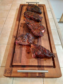 Four barbecued beef ribs with a shiny glaze rest on a BBQ Boards® Brisket BBQ Board® 37" x 12" cutting board with handles, placed on a tiled kitchen counter.