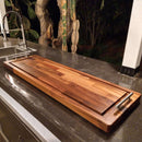 The Brisket BBQ Board® 37" x 12" Cutting Board by BBQ Boards® with metal handles rests on a dark countertop near a modern sink, while trees and plants are visible outside at night.