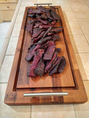 Sliced grilled or smoked meat, likely beef jerky, arranged on the BBQ Boards® Brisket BBQ Board® 37" x 12" cutting board with handles, set on a tiled kitchen countertop.
