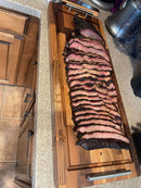 Sliced smoked brisket with a seasoned bark and pink smoke ring, arranged on a BBQ Boards® Brisket BBQ Board® 37" x 12" cutting board on a kitchen counter.