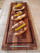 Three pulled pork sandwiches with pickle spears are arranged in a row on a BBQ Boards® Brisket BBQ Board® 37" x 12" cutting board, set atop a tiled kitchen counter.