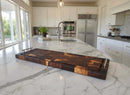 A modern kitchen with white cabinets, stainless steel fridge, built-in ovens, and a marble island featuring the BBQ Boards® Burly BBQ Board® 35 x 12 Cutting Board and a potted plant near the sink. Sunlight streams through large windows.