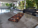 A spacious outdoor kitchen features a sleek concrete countertop, the Burly BBQ Board® 35 x 12 Cutting Board by BBQ Boards® with metal handles, a grill, and lush greenery. Sunlight filters in, making the space bright and inviting.
