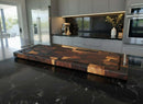 The Burly BBQ Board® 35 x 12 Cutting Board by BBQ Boards® with varied wood tones rests on a black marble island in a modern kitchen featuring grey cabinets and stainless steel appliances.
