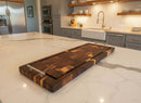 A modern kitchen with a marble countertop showcases the Burly BBQ Board® 35 x 12 Cutting Board by BBQ Boards®, featuring metal handles. Gray cabinets, open shelves, a farmhouse sink, and stainless steel appliances are in the background.