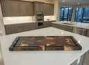 The Burly BBQ Board® 35 x 12 Cutting Board by BBQ Boards® with metal handles rests on a white kitchen island in a modern kitchen with gray cabinets and stainless steel appliances.