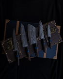 The Folded Steel Dahlia 5-Piece Knife Set, made with AUS10 stainless Damascus steel and dark wooden handles, is arranged on a closed vintage-style brown suitcase with brass hardware against a dark backdrop.