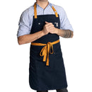 A person in a light blue shirt and Folded Steel Chef Apron with Eco-Friendly rPET Canvas, featuring tan straps and a front pocket, stands with hands clasped. Visible arm tattoos; plain white background evokes a sustainable kitchen vibe.