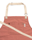 The Folded Steel Chef Apron features eco-friendly rPET canvas in pink, beige adjustable neck and waist straps, a front pocket with button details, and a white side tag—combining style and sustainability for your kitchen.