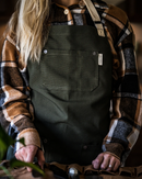 A person wears the Folded Steel Chef Apron with Eco-Friendly rPET Canvas over a brown, black, and white plaid shirt, standing indoors and facing forward with hands resting on a surface. Only the torso and hands are visible.