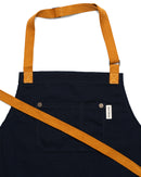 The Folded Steel Chef Apron with Eco-Friendly rPET Canvas features dark blue fabric, a front pocket with a white tag, brass rivets, and adjustable orange straps—a stylish and sustainable kitchen essential.